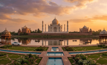 A wide-angle, symmetrical view of the Taj Mahal at sunset, viewed from across the Yamuna River. The white marble mausoleum and its minarets are perfectly reflected in the water, framed by manicured Mughal gardens and a clear, golden-hued sky.