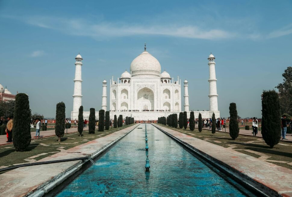 "The Taj Mahal in Agra, India, with its white marble dome and four minarets reflected in the long central water channel, surrounded by manicured gardens under a clear blue sky
