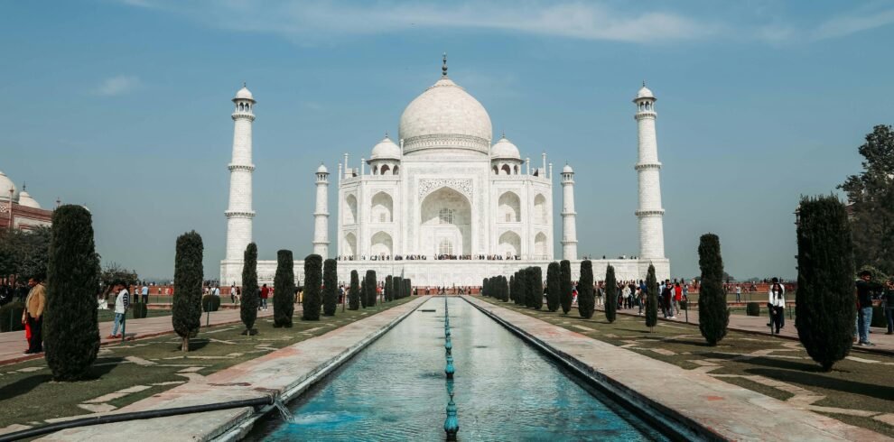 "The Taj Mahal in Agra, India, with its white marble dome and four minarets reflected in the long central water channel, surrounded by manicured gardens under a clear blue sky