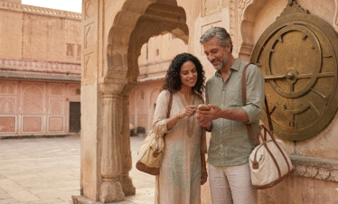 A man and woman smiling while looking at a phone together in a sun-drenched Jaipur palace courtyard. The scene features ornate Rajasthani arches and a large ancient sundial from the Jantar Mantar in the background.