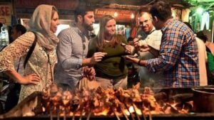 An atmospheric dusk scene near Jama Masjid, focusing on a charcoal grill loaded with skewers of sizzling kebabs, set within a crowded lane.