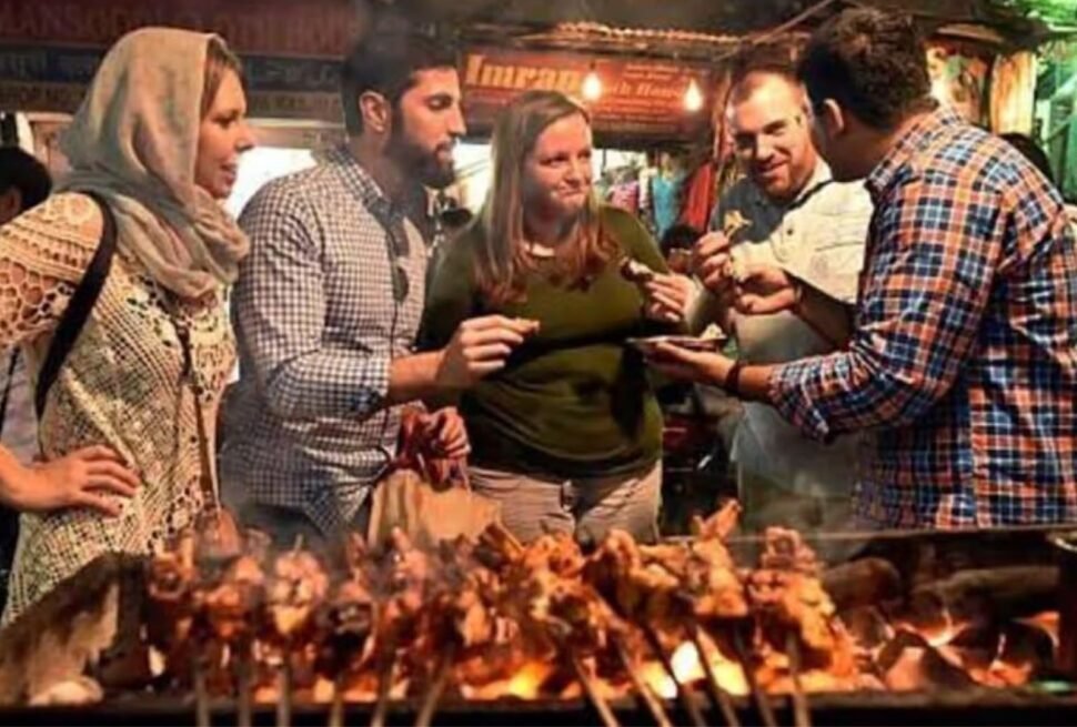 An atmospheric dusk scene near Jama Masjid, focusing on a charcoal grill loaded with skewers of sizzling kebabs, set within a crowded lane.