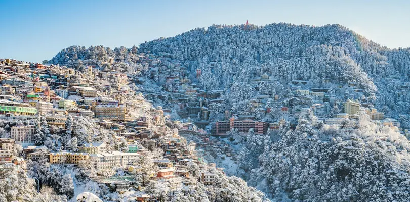 Shimla – The Queen of Hills. Panoramic view of a snow‑covered hillside town with colorful buildings cascading down the mountain slopes, surrounded by forested hills blanketed in snow under a bright blue sky.