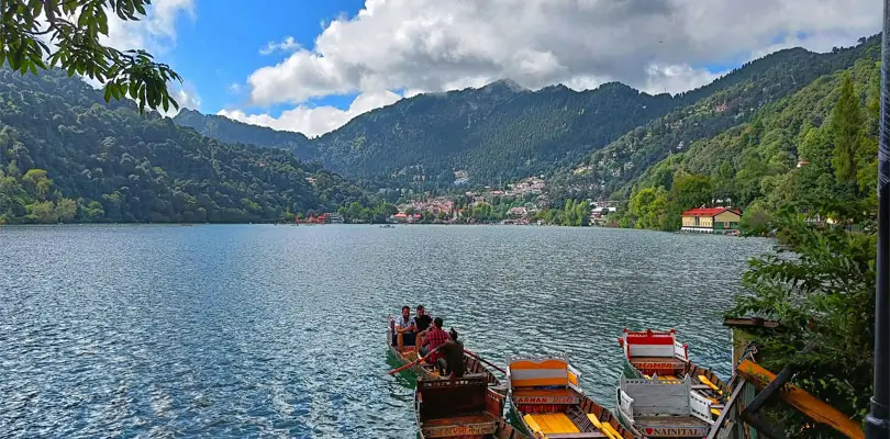 Nainital – The City of Lakes Colorful wooden boats docked along a calm lake in Nainital, surrounded by lush green hills and mountains, with buildings nestled among trees under a partly cloudy sky.