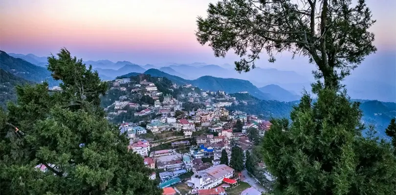 Mussoorie – Weekend Escape Scenic view of a hill station town built on mountain slopes, surrounded by lush green pine trees and layered ranges fading into the horizon under a pastel sunrise sky.