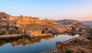 A scenic daytime shot of Amer Fort showing the expansive ramparts and the winding cobblestone path leading up the hill. The fort's majestic structures overlook Maota Lake and the surrounding Kesar Kyari gardens, capturing the grand scale of the UNESCO World Heritage site.