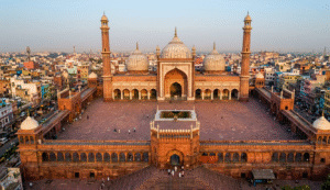 An expansive aerial wide-angle shot of the Jama Masjid in Delhi at sunrise. The massive red sandstone courtyard is nearly empty, highlighting the symmetry of the three white marble domes and two towering minarets against a backdrop of the densely packed Old Delhi cityscape.