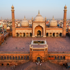 An expansive aerial wide-angle shot of the Jama Masjid in Delhi at sunrise. The massive red sandstone courtyard is nearly empty, highlighting the symmetry of the three white marble domes and two towering minarets against a backdrop of the densely packed Old Delhi cityscape.