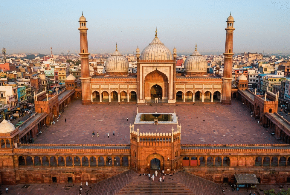 An expansive aerial wide-angle shot of the Jama Masjid in Delhi at sunrise. The massive red sandstone courtyard is nearly empty, highlighting the symmetry of the three white marble domes and two towering minarets against a backdrop of the densely packed Old Delhi cityscape.
